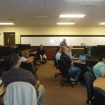 An office meeting in progress with attendees seated at tables looking towards a speaker at the front of the room.