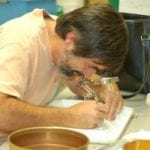 A man with a beard using a microscope to examine or work on a small object at a cluttered workstation.