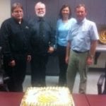 Four people standing around a table with a large celebratory cake in an office setting.