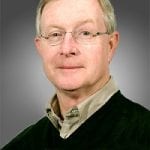 Portrait of an older man with light skin and gray hair, wearing glasses and a dark green sweater over a beige shirt against a gray background.