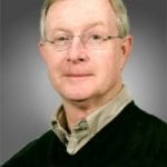 Portrait of an elderly man with glasses, wearing a green sweater over a collared shirt against a gray background.