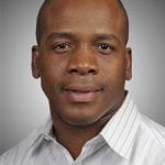 Portrait of a bald black man wearing a white striped shirt against a gray background. he is looking directly at the camera.