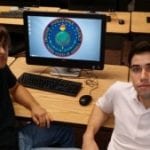 Two men sitting at a desk with a computer screen displaying a colorful circular graphic between them.