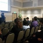 Audience listening to a presenter in a conference hall with rows of chairs and a large projection screen.