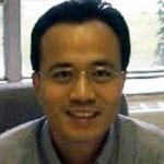 A smiling asian man wearing glasses and a gray polo shirt, seated indoors with a window in the background.