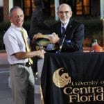 Two men shaking hands at a university of central florida event, smiling and holding a trophy with the university's logo on the table in front of them.