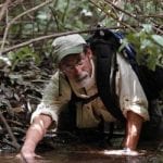 A man with a beard, wearing a hat and glasses, crawls through muddy water under dense forest brush.