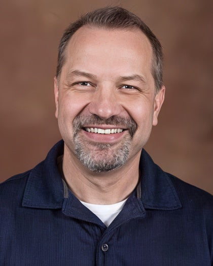 A middle-aged man with short hair and a goatee, wearing a navy collared shirt, smiles at the camera against a brown background.