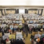 Overhead view of a busy conference hall with attendees viewing numerous research posters on display boards.