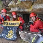 Three smiling people in red helmets and outdoor gear holding a 