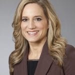 Professional headshot of a smiling woman with long blonde hair, wearing a brown blazer.