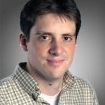 Portrait of a man with dark hair wearing a checkered shirt, smiling slightly against a gray background.