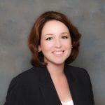 Professional portrait of a smiling woman with shoulder-length brown hair, wearing a black suit jacket against a grey background.