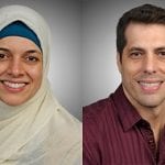 Portrait of a woman wearing a white hijab and a man in a burgundy shirt, both smiling against a grey background.