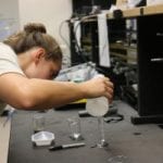 A young woman in a lab pours a substance from a beaker into a flask while other students work in the background.