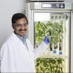 A scientist in a lab coat examines plant samples in a growth chamber, smiling confidently at the camera.