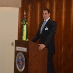 A man in a suit standing at a podium with a microphone, with a brazilian flag on the side, inside a room with wooden panel walls.