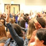 A diverse crowd of people raising their hands enthusiastically in a crowded indoor event.