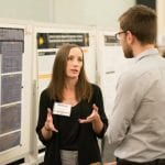 A woman with a name tag gestures while talking to a man at a poster presentation.