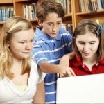 Three teenagers, two girls and a boy, using a laptop together in a library.