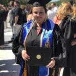 A female graduate in anthropology, dressed in a black cap and gown, wearing honors cords, smiling and holding her diploma during a graduation ceremony.