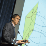 A man in a suit presenting a mathematical lecture beside a projected graph.