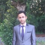 Man in a gray suit and purple tie standing before a tree and green foliage.