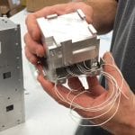 A person holding a small, intricate metal device with multiple wires connected to it, standing beside another metal component on a workbench.