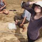 A woman adjusting her hat at an archaeological dig site, with two others working in the background.