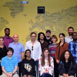 Group of diverse people posing in front of a world map wall mural, smiling in a classroom setting.