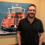 A man in a black polo shirt smiling, standing in front of a framed photograph of a large red ship on the ocean.