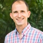 A man with a shaved head smiling outdoors, wearing a plaid shirt with a blurred green foliage background.
