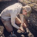 A person in a plaid shirt and cap kneels while carefully excavating an artifact from the soil with a small trowel.