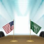Two national flags, usa and saudi arabia, standing on a conference table in a room with a blurred, blue-lit background.