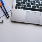 Laptop with earphones, notepad, and pens on a white desk, representing a typical workspace setup.