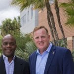 Two men smiling and standing outside a building with palm trees in the background. one man is bald, the other has reddish hair.