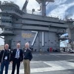 Three men stand in front of an aircraft carrier with a 