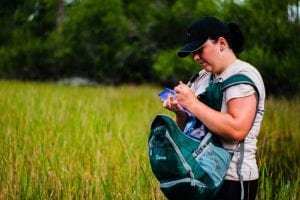 A biologist records data in a notebook while standing in a wetland, wearing a vest and carrying a backpack.