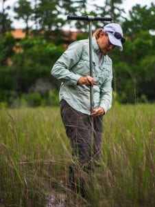 A woman in a green shirt and tan pants examining soil samples using a t-shaped soil probe in a grassy field.