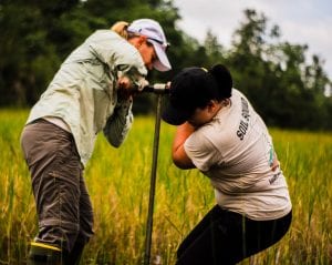 Two women in casual outdoor attire are examining soil samples in a grassy field using a soil auger.