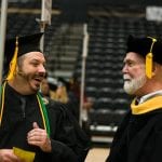 A young male graduate in a cap and gown laughing while talking to an older male professor, also in graduation regalia, in a dimly lit auditorium.