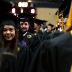 Graduates in caps and gowns standing in line at a graduation ceremony, with a focus on a smiling young woman looking towards the camera.