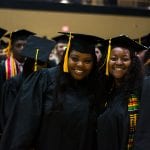 Two smiling graduates in black gowns and caps, one wearing a multicolored stole, standing together during a graduation ceremony.