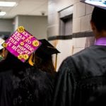 Graduates walking in a corridor, one wearing a decorated cap with text "kick some grad a$$!" and sunflower designs.