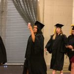 Graduation procession in a gymnasium with a female graduate in the center taking a selfie.