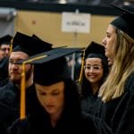 Graduates in black caps and gowns at a commencement ceremony, with some looking around and others focused forward.