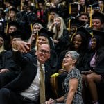 Man in a suit and a woman smiling as they take a selfie with a group of diverse graduating students in caps and gowns in the background.