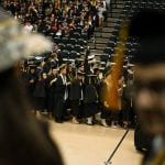 Graduates in black robes and caps line up at a graduation ceremony, with a focus on one smiling graduate through a blurred foreground.