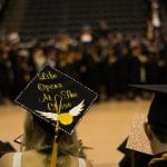 A graduate wearing a decorated cap that reads "life opens at the close" at a crowded graduation ceremony.