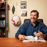 Man smiling at his desk in an office, surrounded by books and cultural artifacts, flipping through a book.